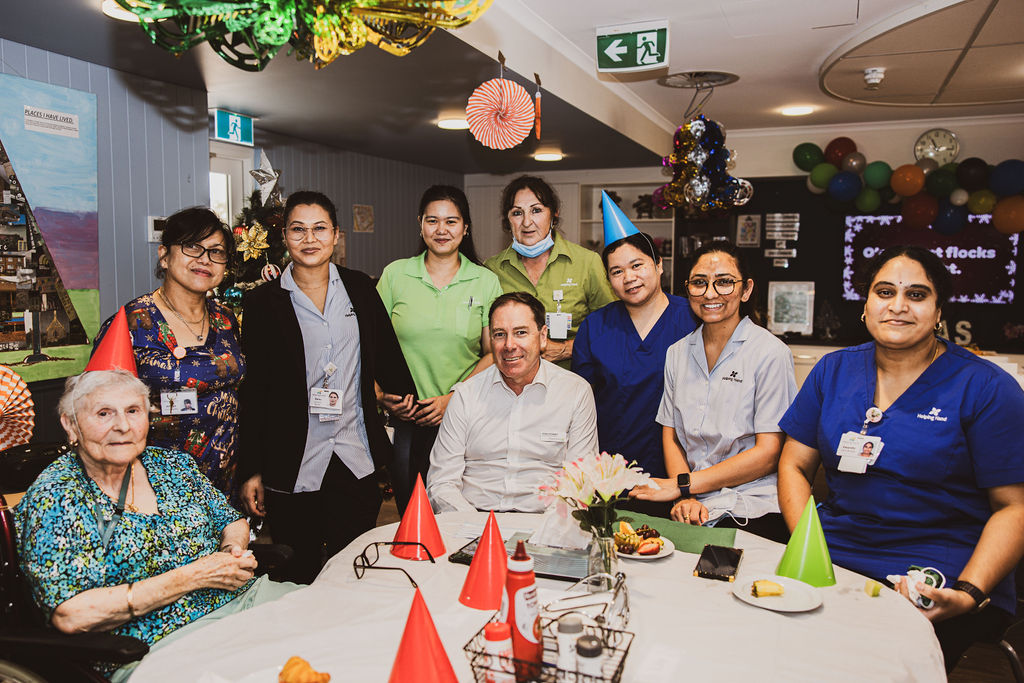 A group of employees stand around a table celebrating a special occasion. There are part decorations and sweets. They are smiling at the camera.