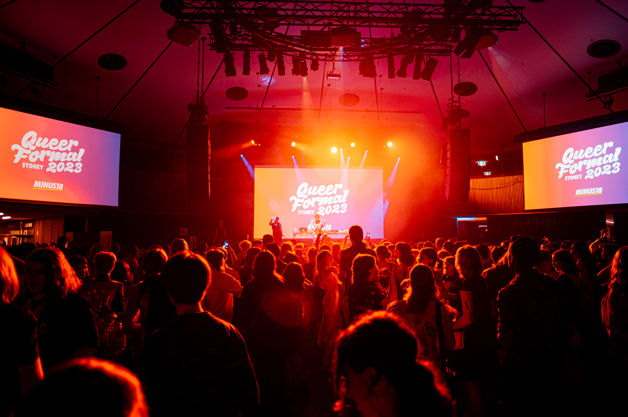 Queer Formal 2025, a large room filled with coloured lights and young people dancing. The words "Queer Formal" are projected onto a screen in the background.