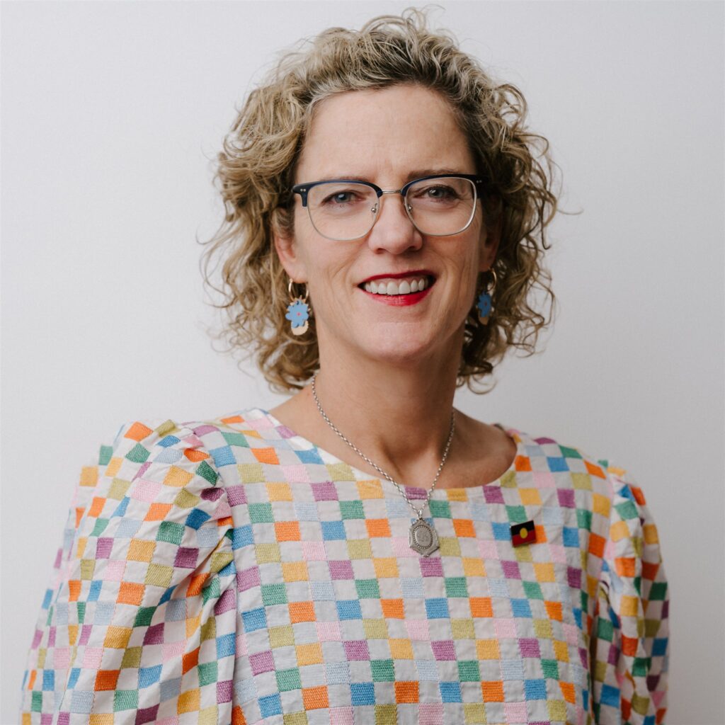 Headshot of Patty Kinnersly. Patty has curly, short blonde hair and wears glasses, pendant earrings and a colourful blouse.