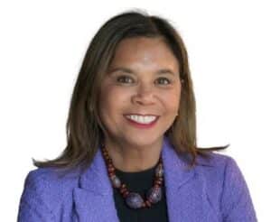 Women wearing a purple blazer, black shirt, a beaded necklace and has brown hair.
