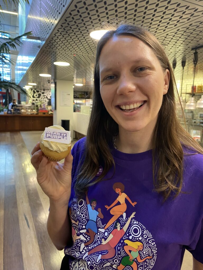 Brenna Harding, a person with long brown hair wearing a purple t-shirt holding a Wear it Purple cupcake