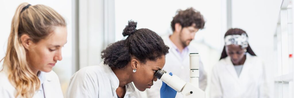 A woman scientist looking into a microscope surrounded by other scientists who are taking notes.