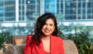 Sunita Gloster sitting in a grey chair in an office setting. She has long, curly, dark hair, brown skin and is wearing an orange business suit and is smiling at the camera.