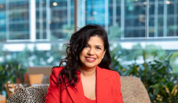 Sunita Gloster sitting in a grey chair in an office setting. She has long, curly, dark hair, brown skin and is wearing an orange business suit and is smiling at the camera.