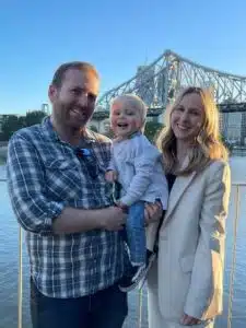 A man in a checkered shirt holding a baby next to a woman in a beige blazer.